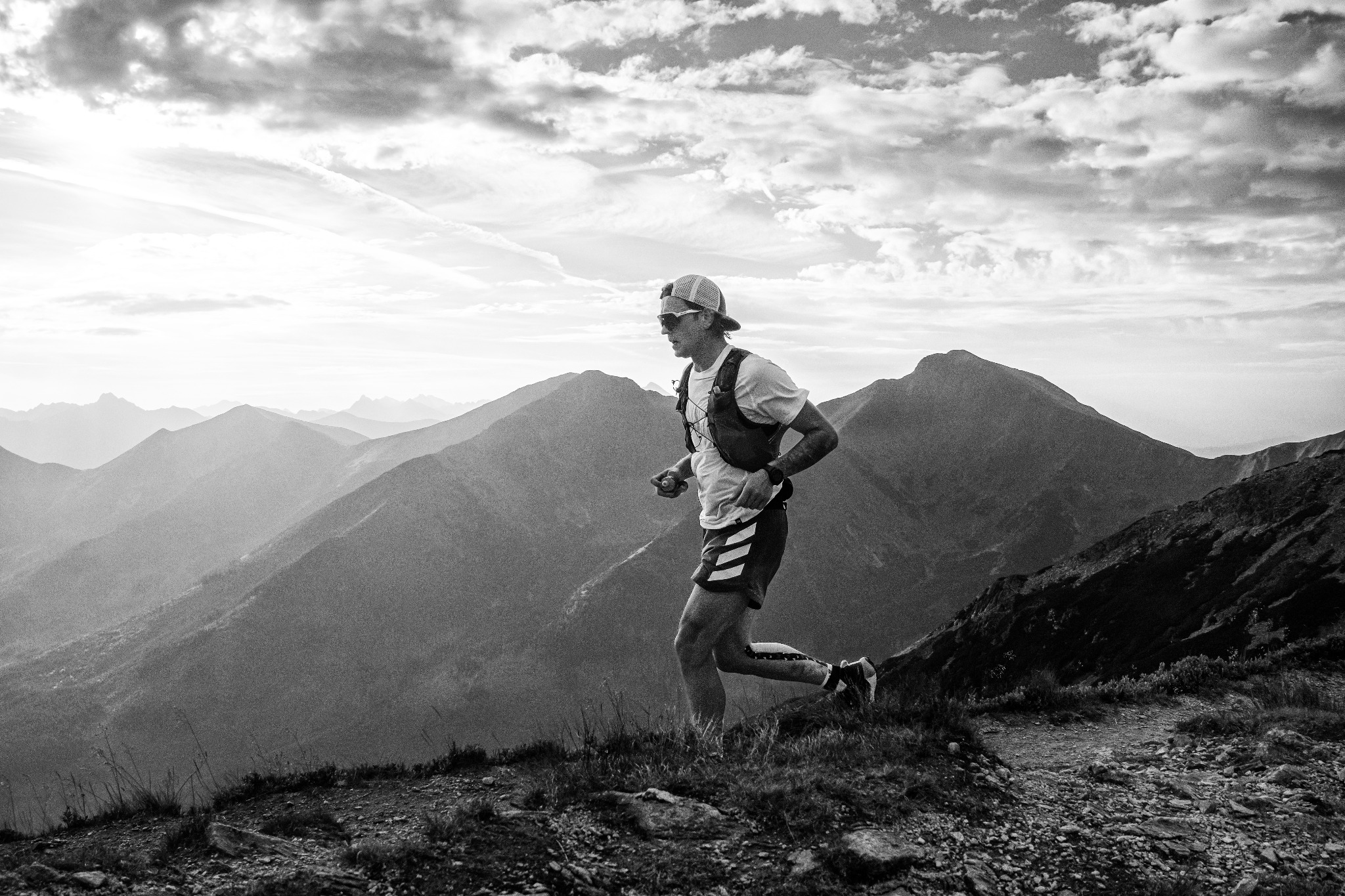 Czarno-białe zdjęcie biegacza górskiego z plecakiem, w trakcie biegu na tle gór i pochmurnego nieba. Widok z profilu, ostre światło zza gór.