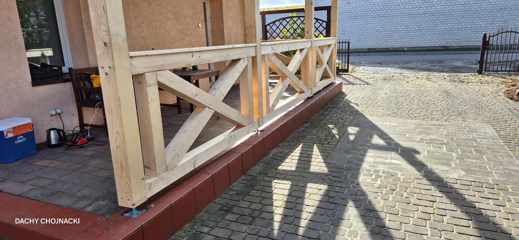 Drewniana konstrukcja z krzyżowymi belkami, prawdopodobnie weranda lub altana, w trakcie budowy. Widoczne elementy mocujące i podłoże z kostki brukowej.