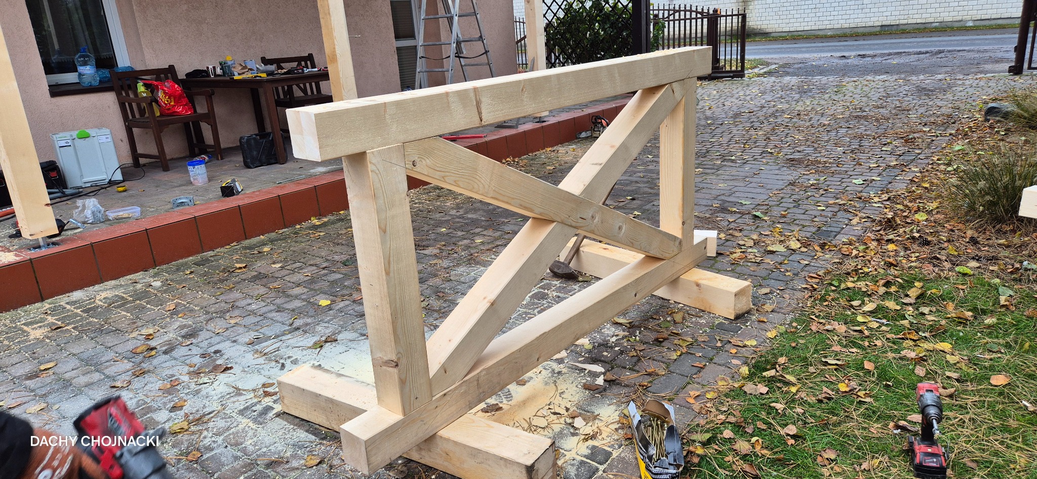 Drewniana konstrukcja szkieletowa, prawdopodobnie fragment dachu, w trakcie montażu na zewnątrz, z widocznymi narzędziami i elementami otoczenia.