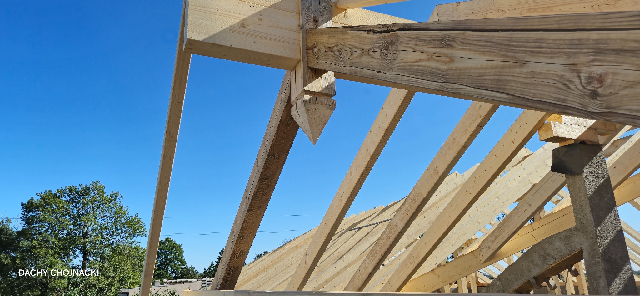 Jasna, drewniana konstrukcja dachu w trakcie budowy, widok z dołu na tle błękitnego nieba. Elementy więźby dachowej z widocznymi detalami łączeń.