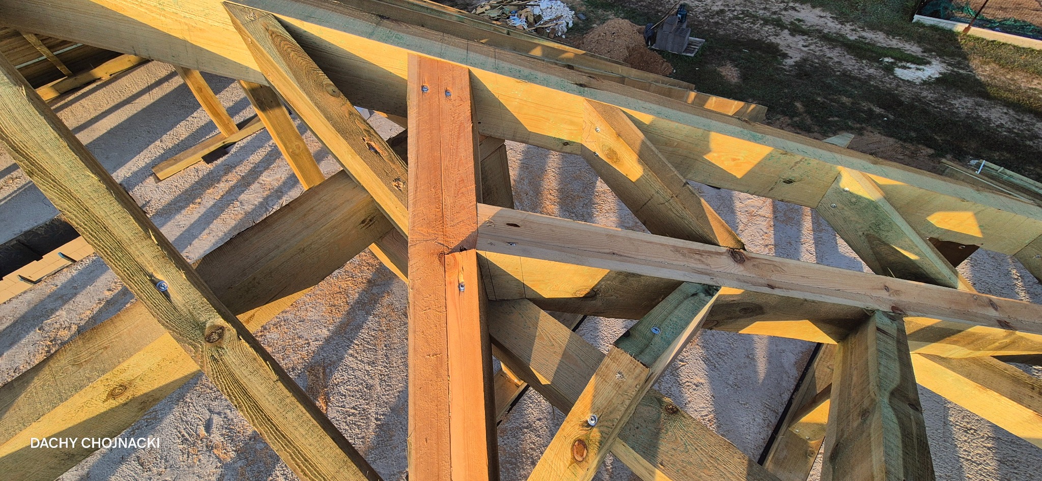 Fragment drewnianej konstrukcji dachu w ciepłym świetle słonecznym, widok z góry, z widocznym podkładem i elementami mocującymi. Dach Chojnacki w rogu.