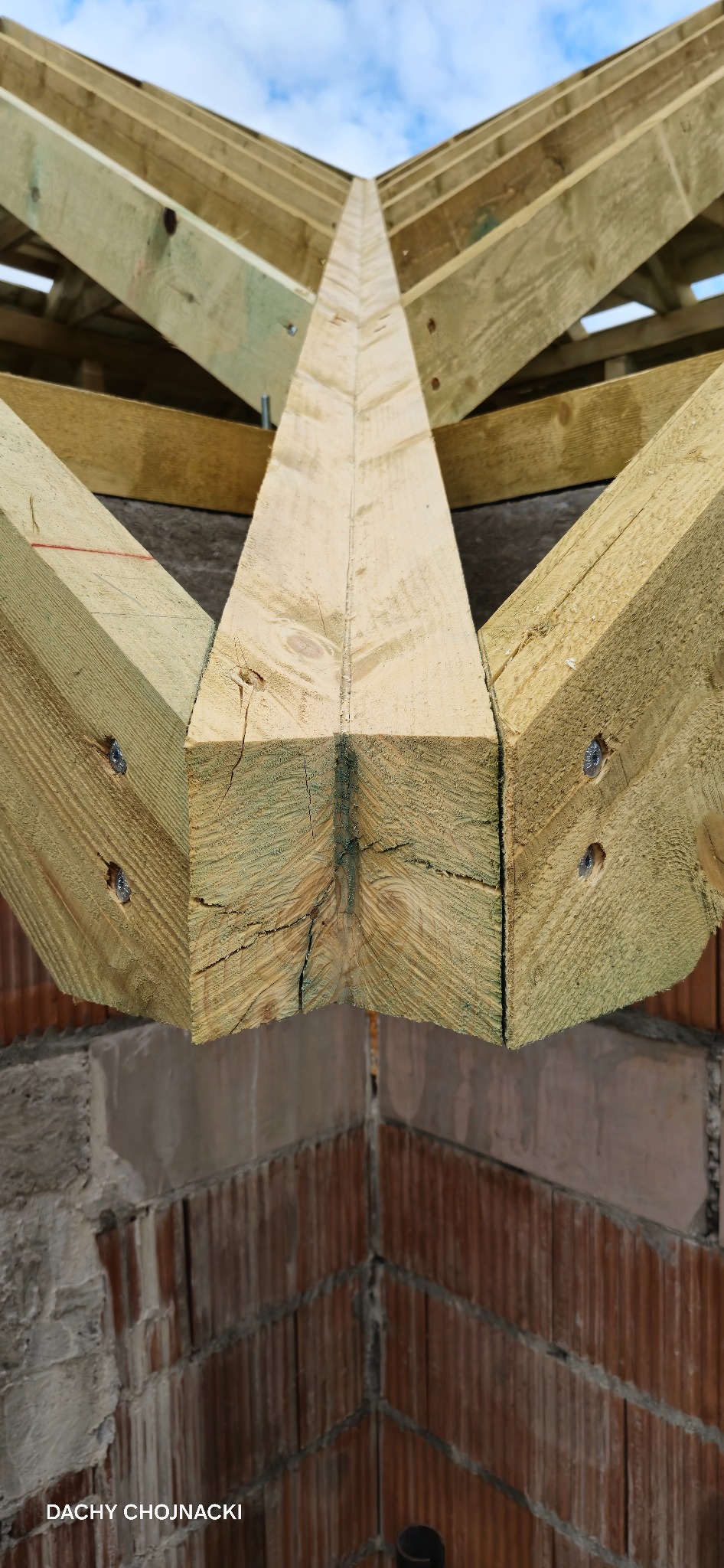 Fragment konstrukcji drewnianej dachu z widocznymi elementami łączeń i śrubami, na tle muru z cegły i fragmentu nieba. Widoczne pęknięcia na drewnie.