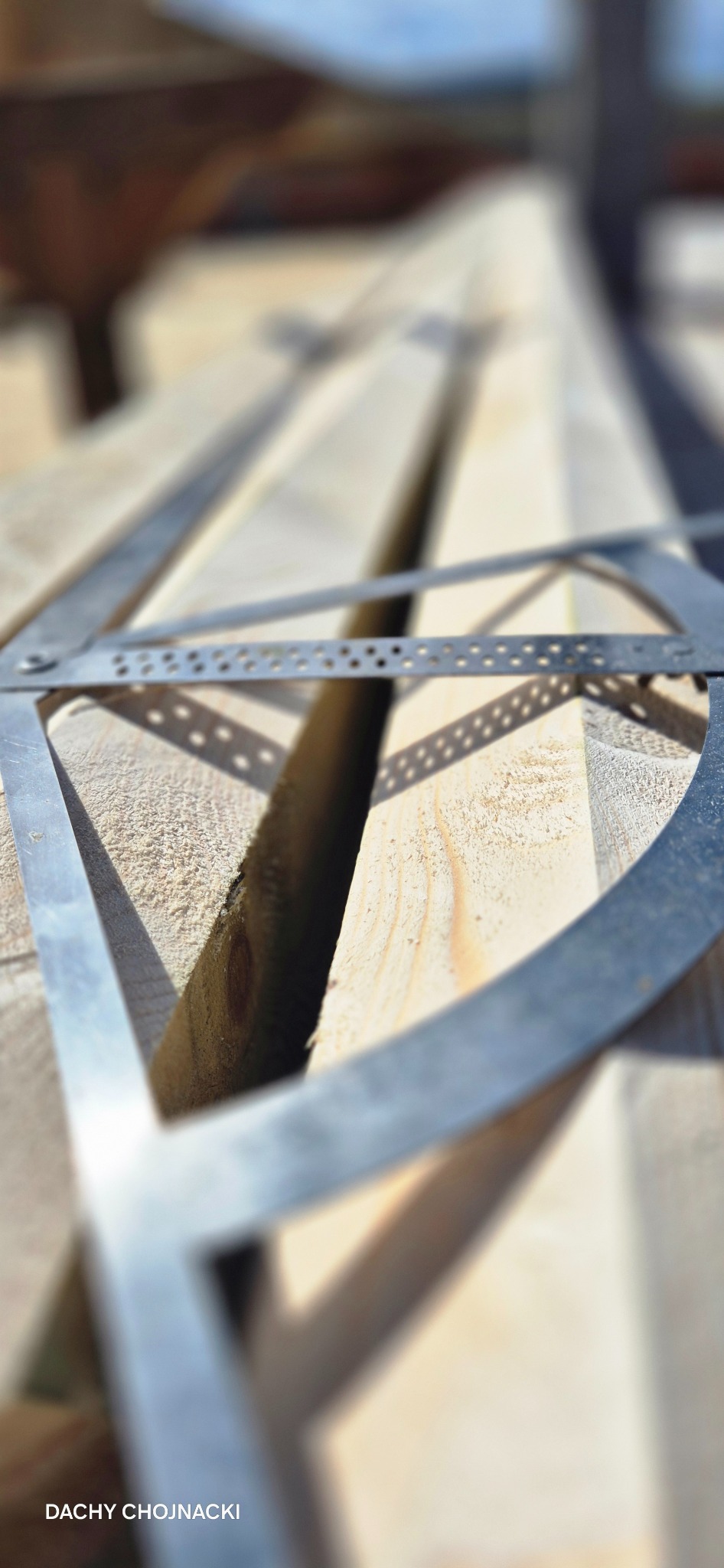 Elementy więźby dachowej z bliska, z metalowym narzędziem pomiarowym, w jasnym oświetleniu. Widoczne ślady obróbki drewna i logo firmy w rogu.