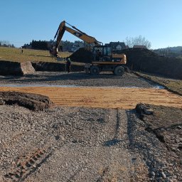 Bud-Max s.c. - Żółta koparka CAT M316D na placu budowy w słoneczny dzień, obok pracownik w zielonym kasku, w tle hałda ziemi i zabudowa jednorodzinna.