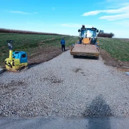 Żółto-niebieska ubijarka obok świeżo wysypanej drogi z kruszywa, w tle koparko-ładowarka i sylwetka mężczyzny na tle pola i błękitnego nieba.
