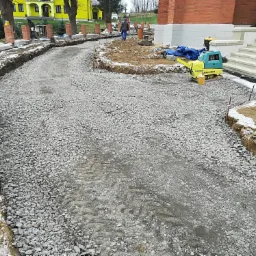 Budowa drogi dojazdowej z podsypką żwirową, ubijarką wibracyjną Ammann i widocznymi wykopami po bokach w zimowej scenerii.