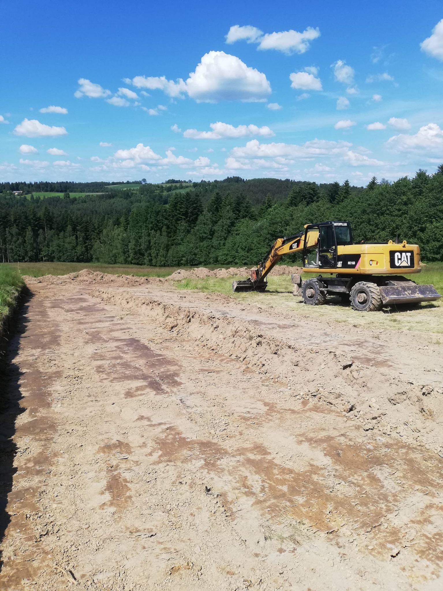 Żółta koparka Caterpillar M316D przygotowuje teren pod drogę dojazdową na tle lasu i błękitnego nieba z chmurami.