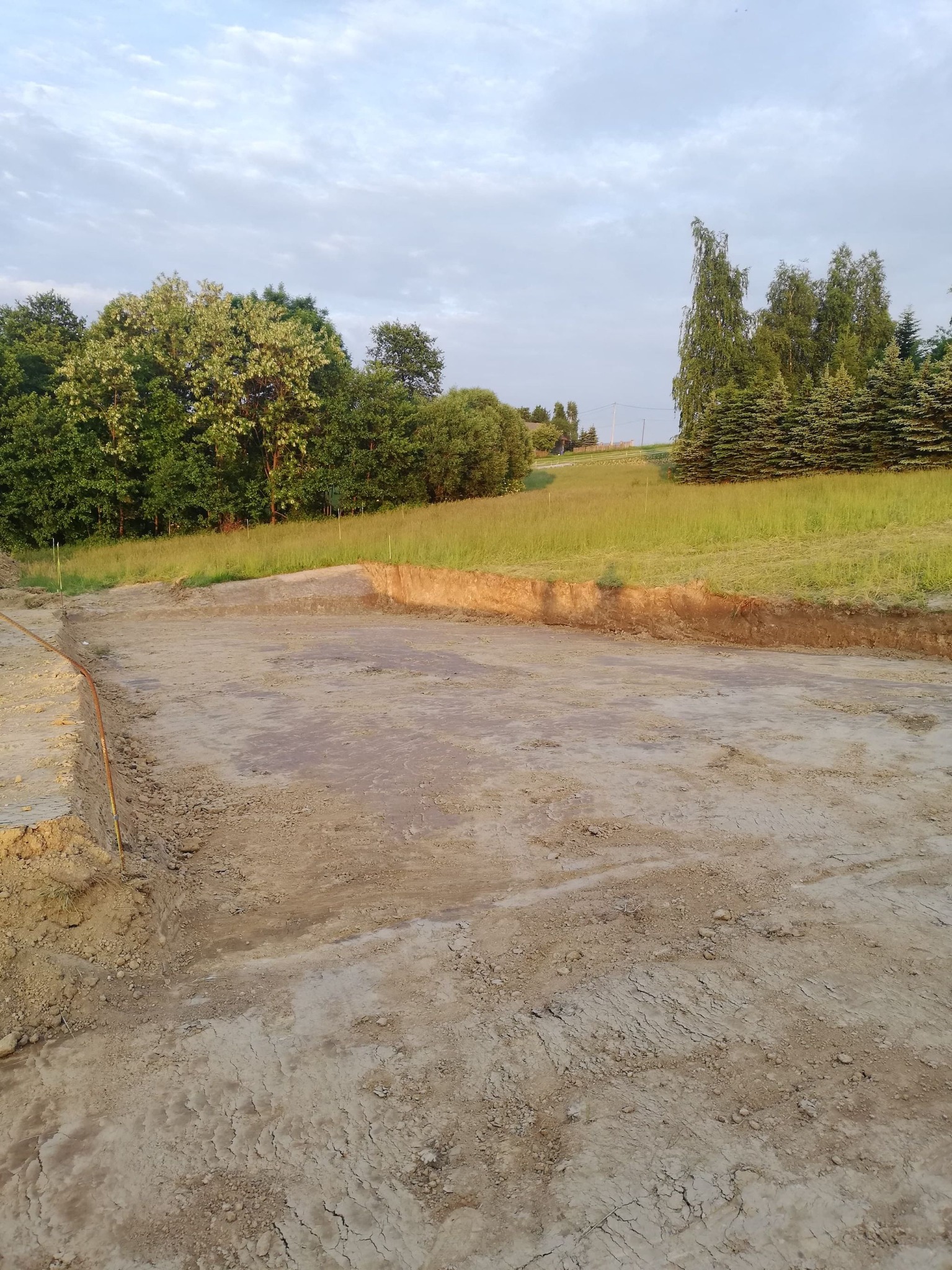 Przygotowany teren pod budowę z widocznym wykopem, grunt wyrównany, w tle drzewa i trawa.