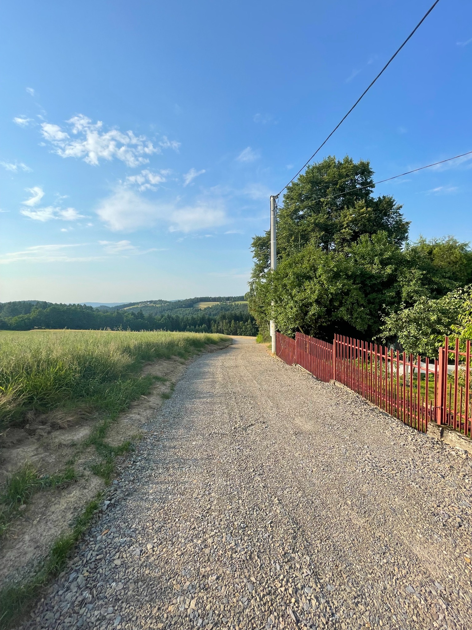 Ukończona droga dojazdowa z tłucznia, prowadząca przez tereny zielone w kierunku zalesionych wzgórz, ograniczona z jednej strony czerwonym metalowym ogrodzeniem.
