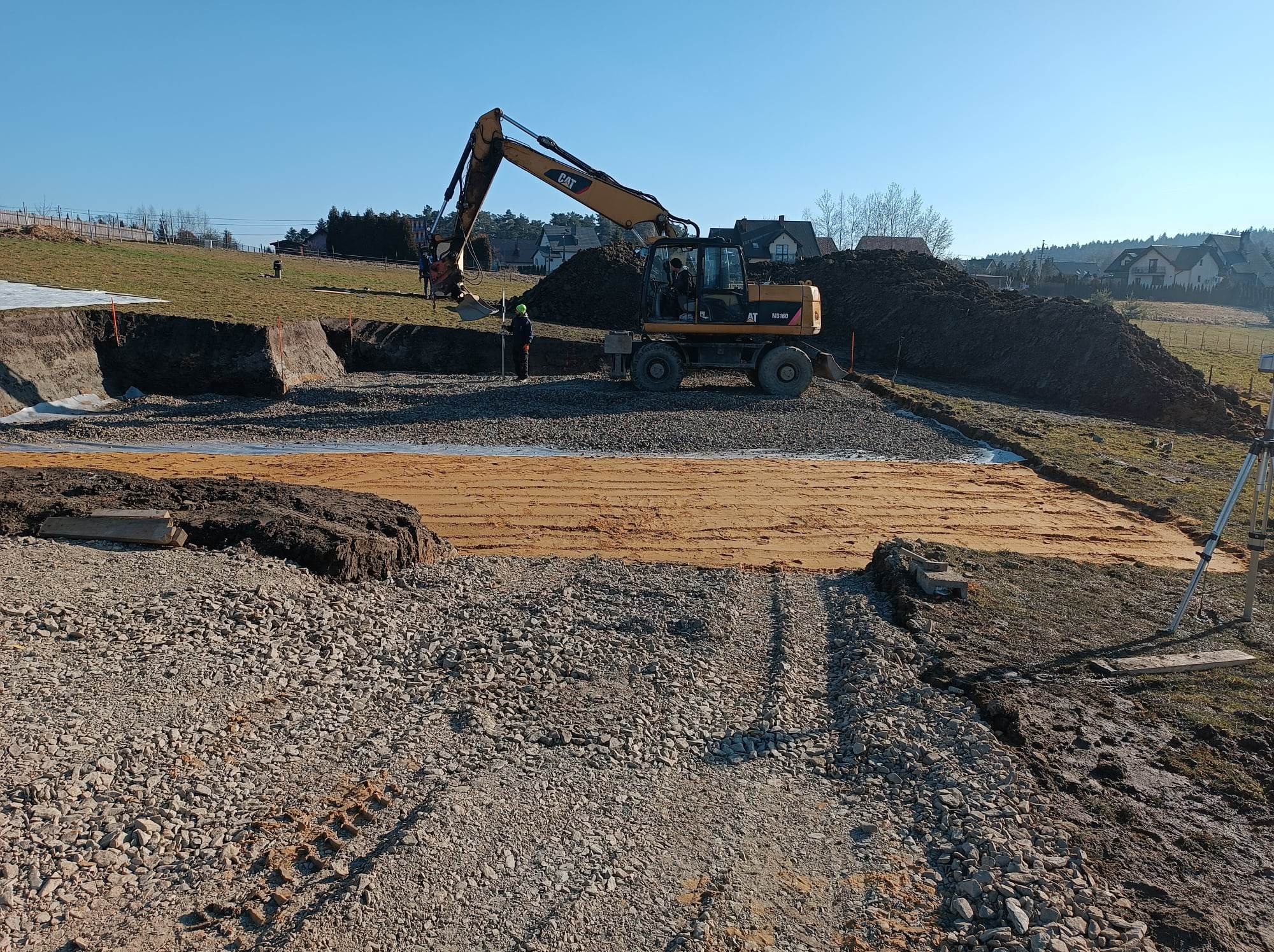 Żółta koparka CAT M316D na placu budowy w słoneczny dzień, obok pracownik w zielonym kasku, w tle hałda ziemi i zabudowa jednorodzinna.