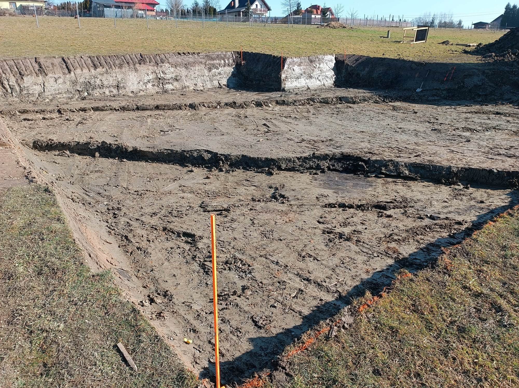 Wykonany wykop pod fundamenty w kształcie litery L, z widocznymi śladami po maszynach, pomarańczowymi znacznikami i tyczkami, oraz warstwami gruntu w przekroju, na tle łąki i zabudowań mieszkalnych.
