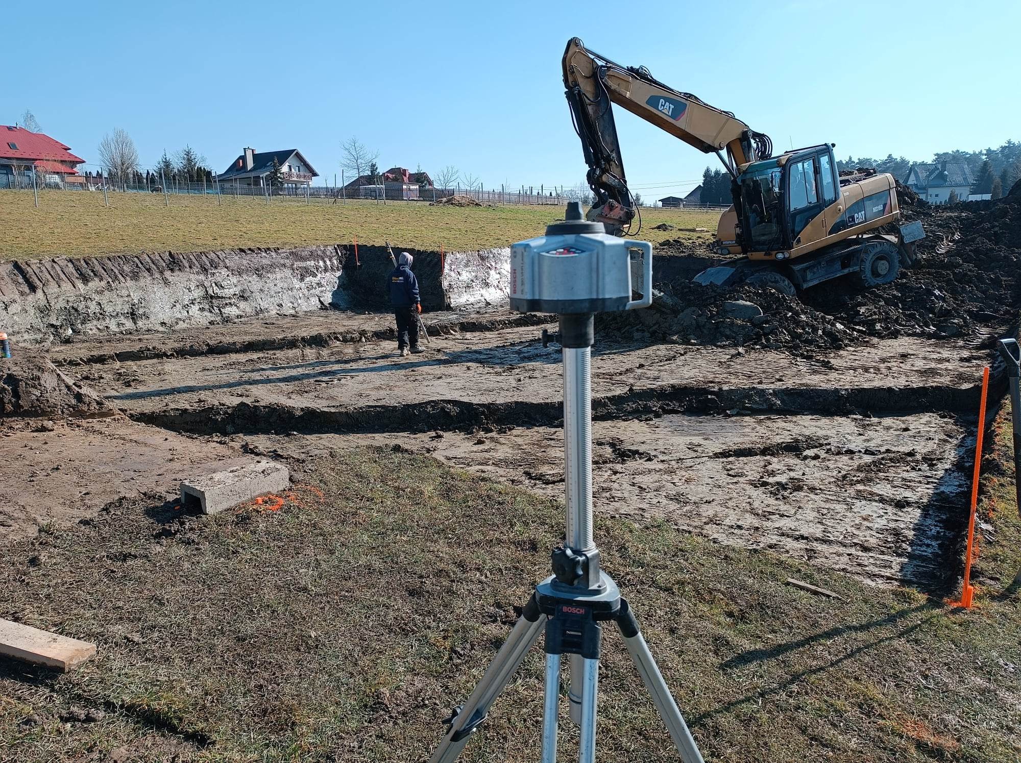 Żółta koparka CAT na placu budowy w Nowym Sączu, widoczny pracownik z tyczką i niwelator laserowy Bosch na statywie, w tle domy.