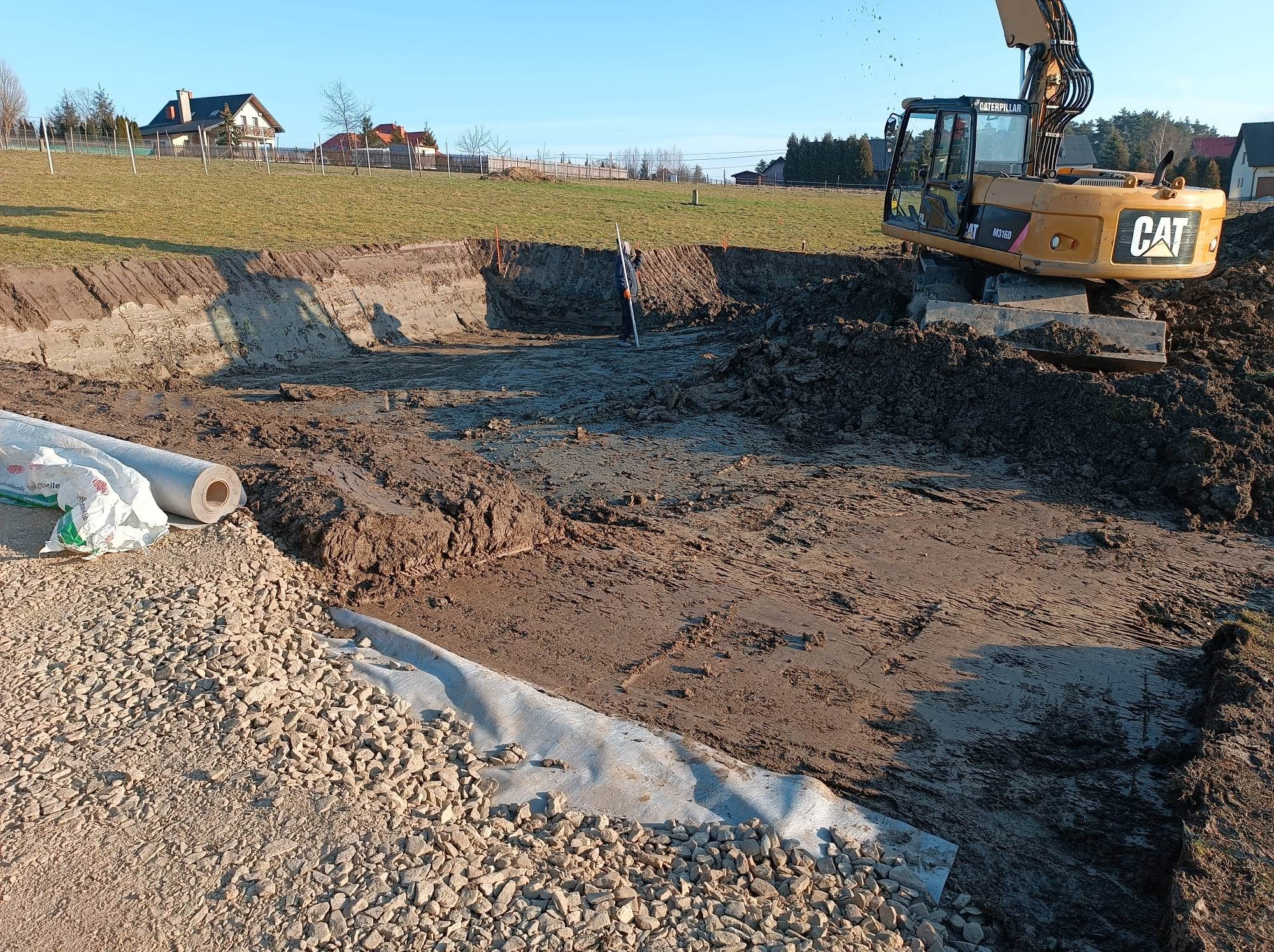 Żółta koparka CAT M316D pracuje przy wykopie, w tle domy, widoczna warstwa geowłókniny i podsypka żwirowa, pracownik z łatą mierniczą kontroluje poziom, wymiana gruntu w Nowym Sączu.
