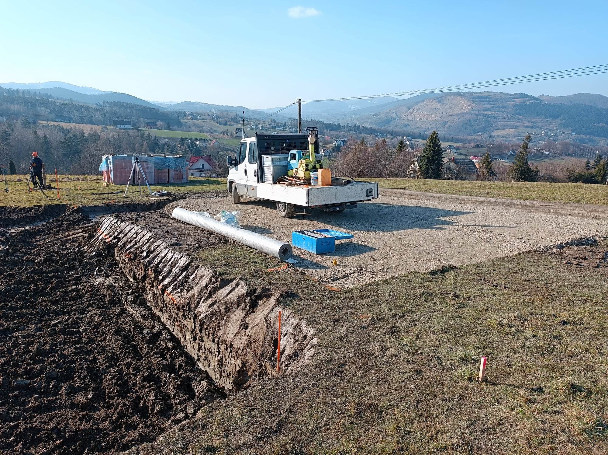 Szeroki wykop w ziemi z widoczną warstwą gliny, przygotowany pod instalację rury drenażowej, Nowy Sącz. W tle biały samochód dostawczy z zagęszczarką i narzędziami, cegły i pracownik z niwelatorem.