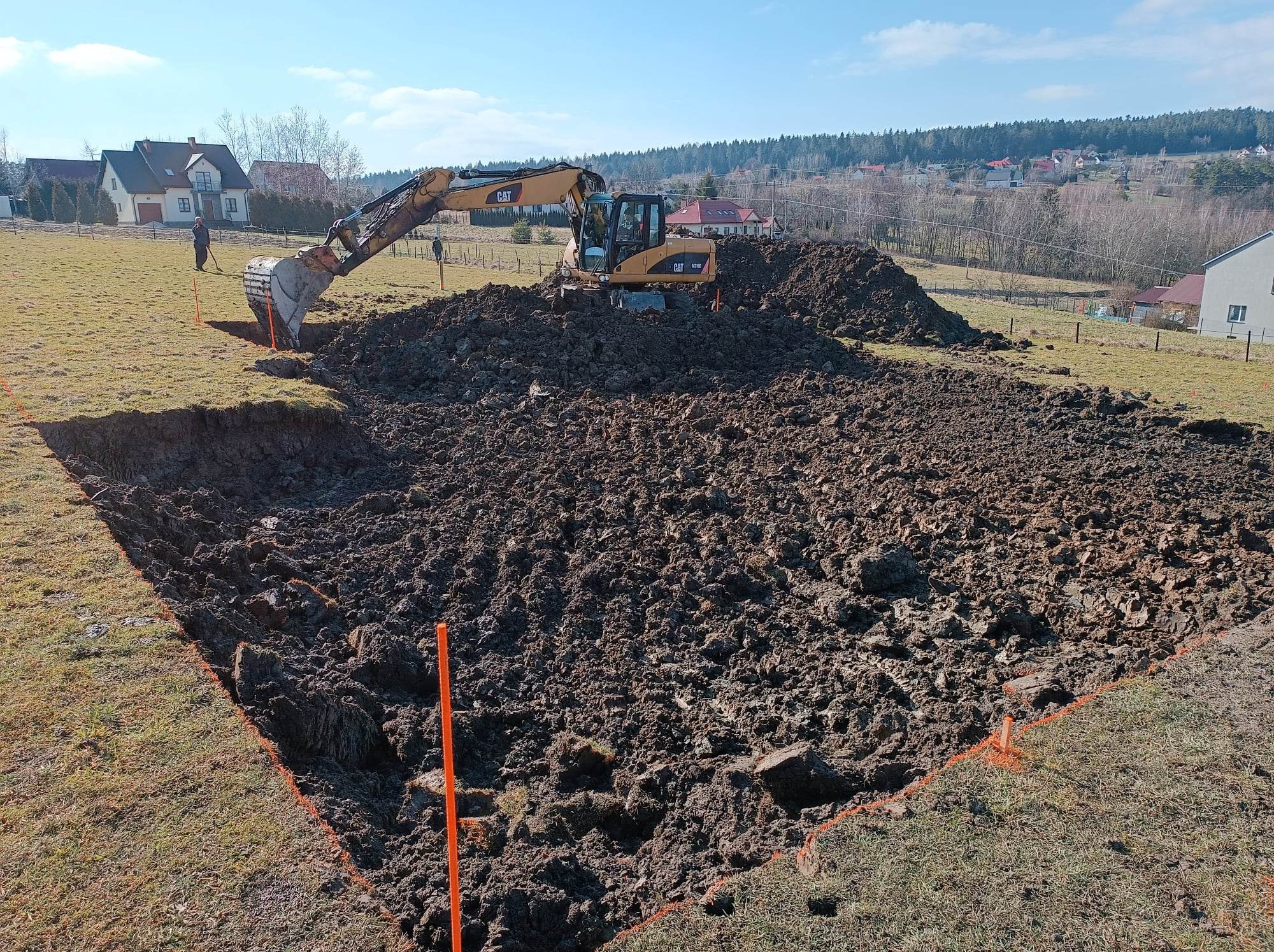 Żółta koparka CAT N316D podczas prac ziemnych na placu budowy w Nowym Sączu, widoczne wyznaczone pomarańczową taśmą granice wykopu i sterty ziemi, w tle zabudowa jednorodzinna i las.