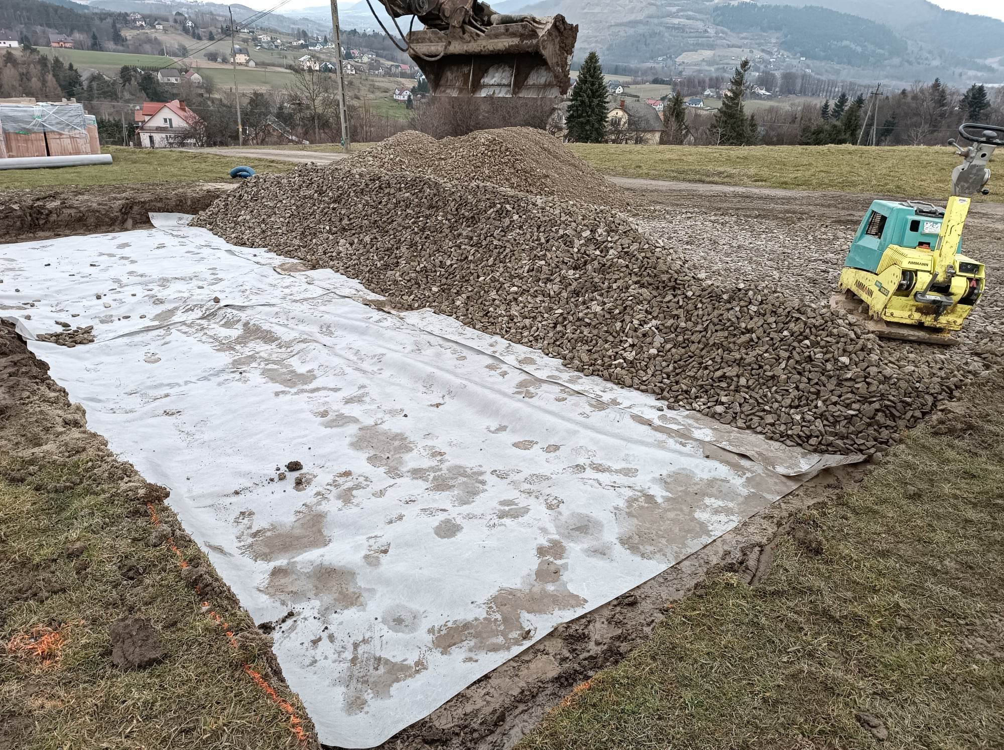Przygotowanie podłoża: wykop wyłożony geowłókniną, obok pryzma żwiru i ubijarka wibracyjna, łyżka koparki w tle, teren pagórkowaty.