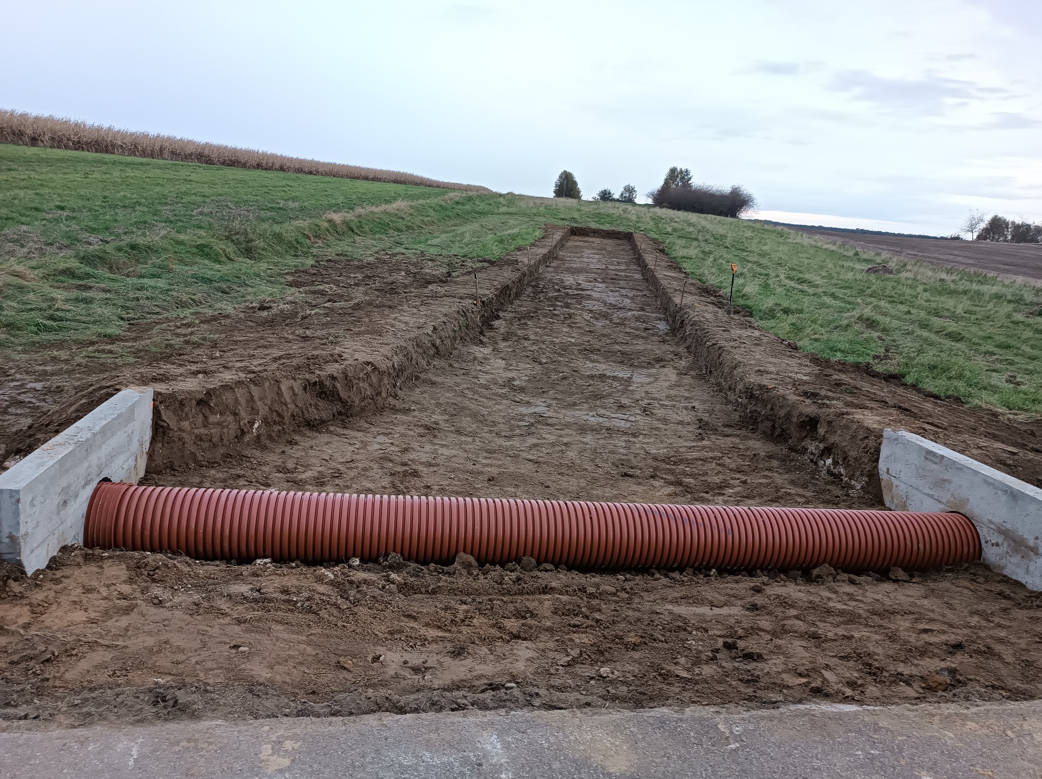Pomarańczowa rura drenażowa ułożona w wykopie z betonowymi ścianami, widok na pole uprawne i pochmurne niebo w tle.