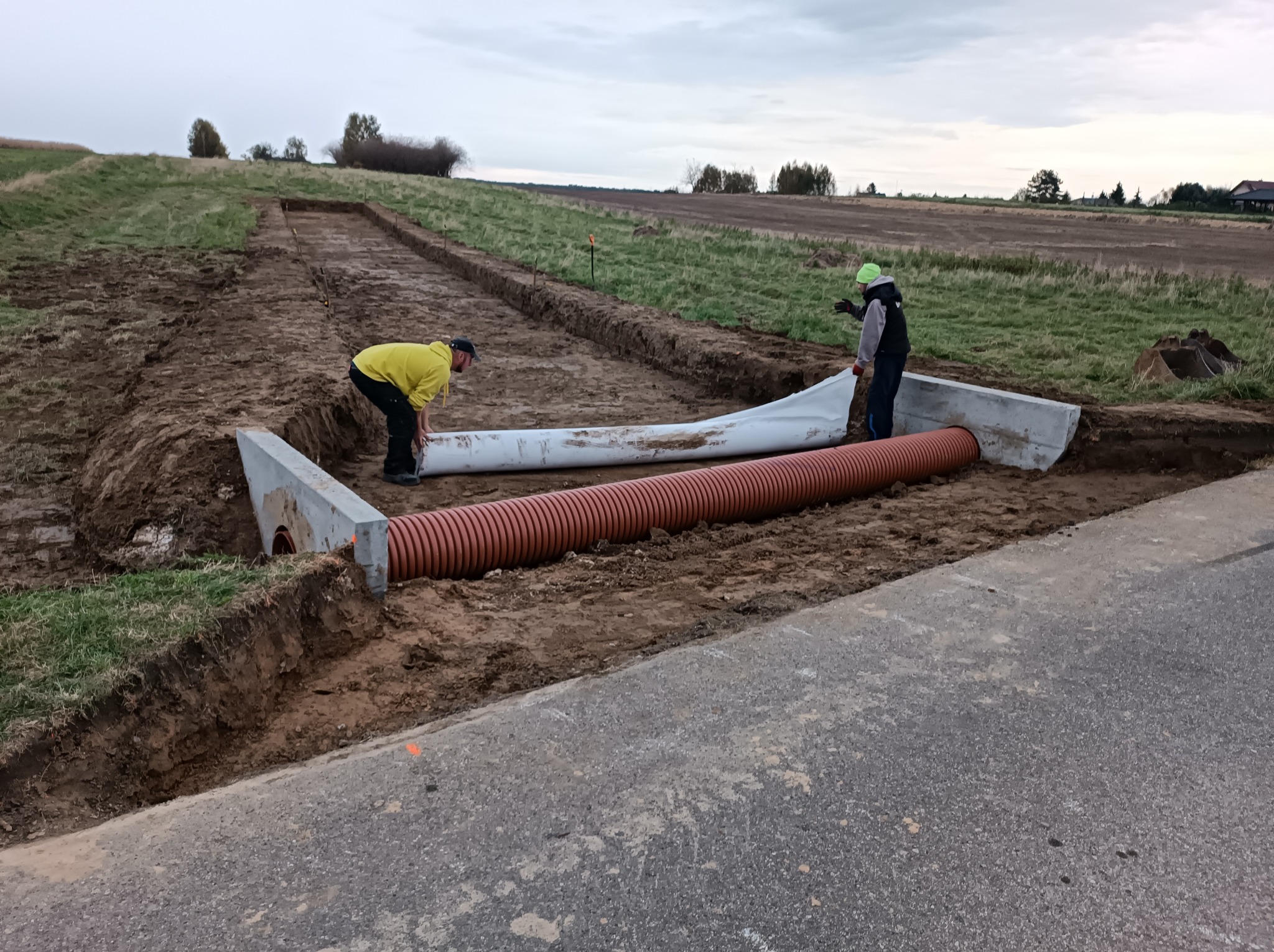Dwóch robotników instaluje pomarańczową rurę drenażową z betonowymi elementami na skraju drogi, widoczne wykopane rowy w ziemi i odległe pole.