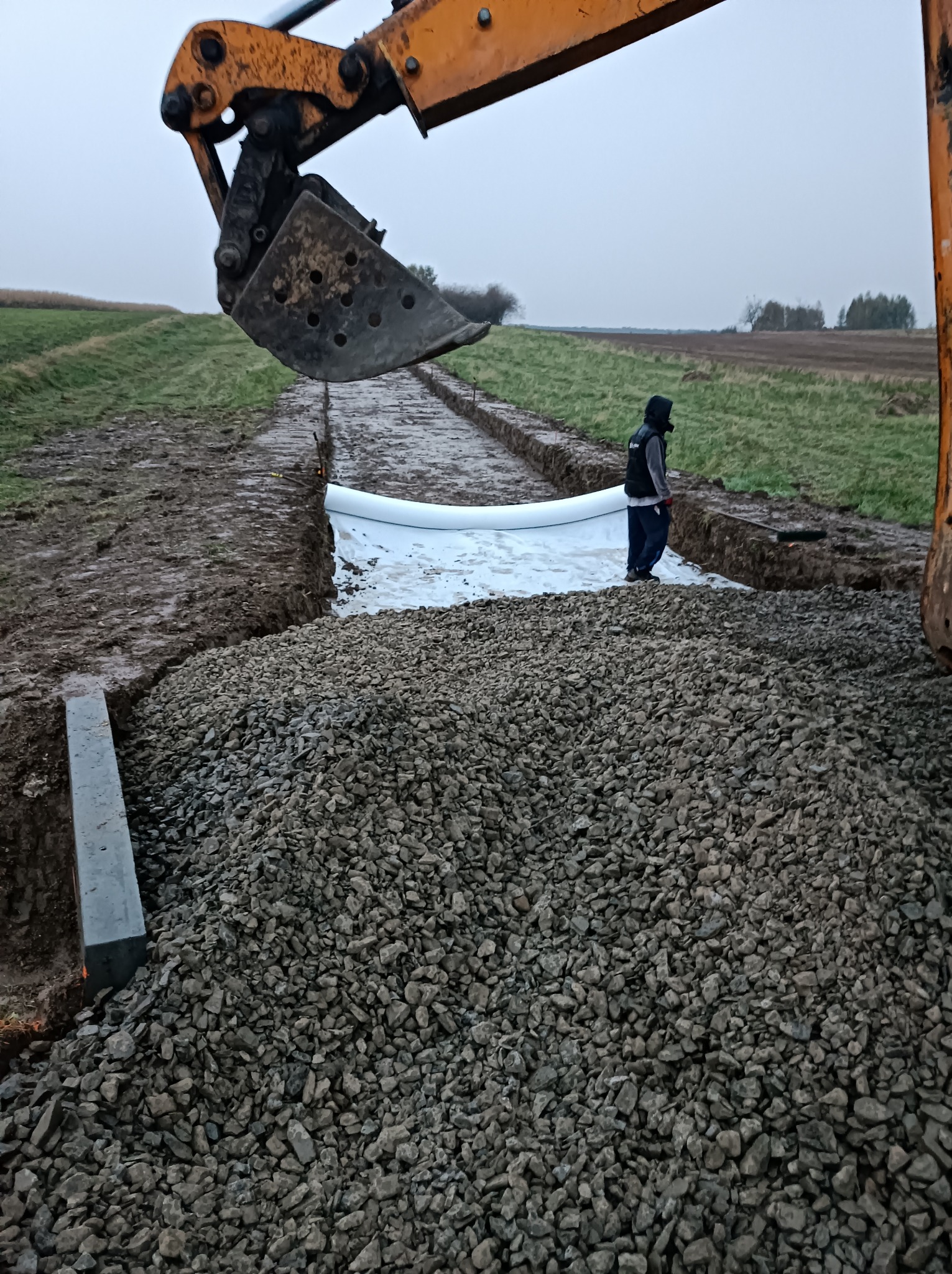 Żwirowa podsypka w wykopie zabezpieczonym białą geowłókniną, z żółtą koparką w tle i pracownikiem w odzieży roboczej, widok z boku na etapie prac.
