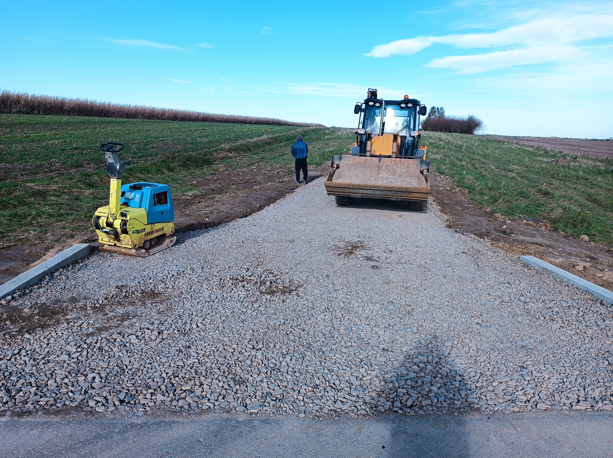 Żółto-niebieska ubijarka obok świeżo wysypanej drogi z kruszywa, w tle koparko-ładowarka i sylwetka mężczyzny na tle pola i błękitnego nieba.
