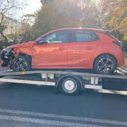 Uszkodzony, pomarańczowy Opel Corsa na platformie lawety, widoczne uszkodzenia przodu pojazdu, jesienne drzewa w tle.