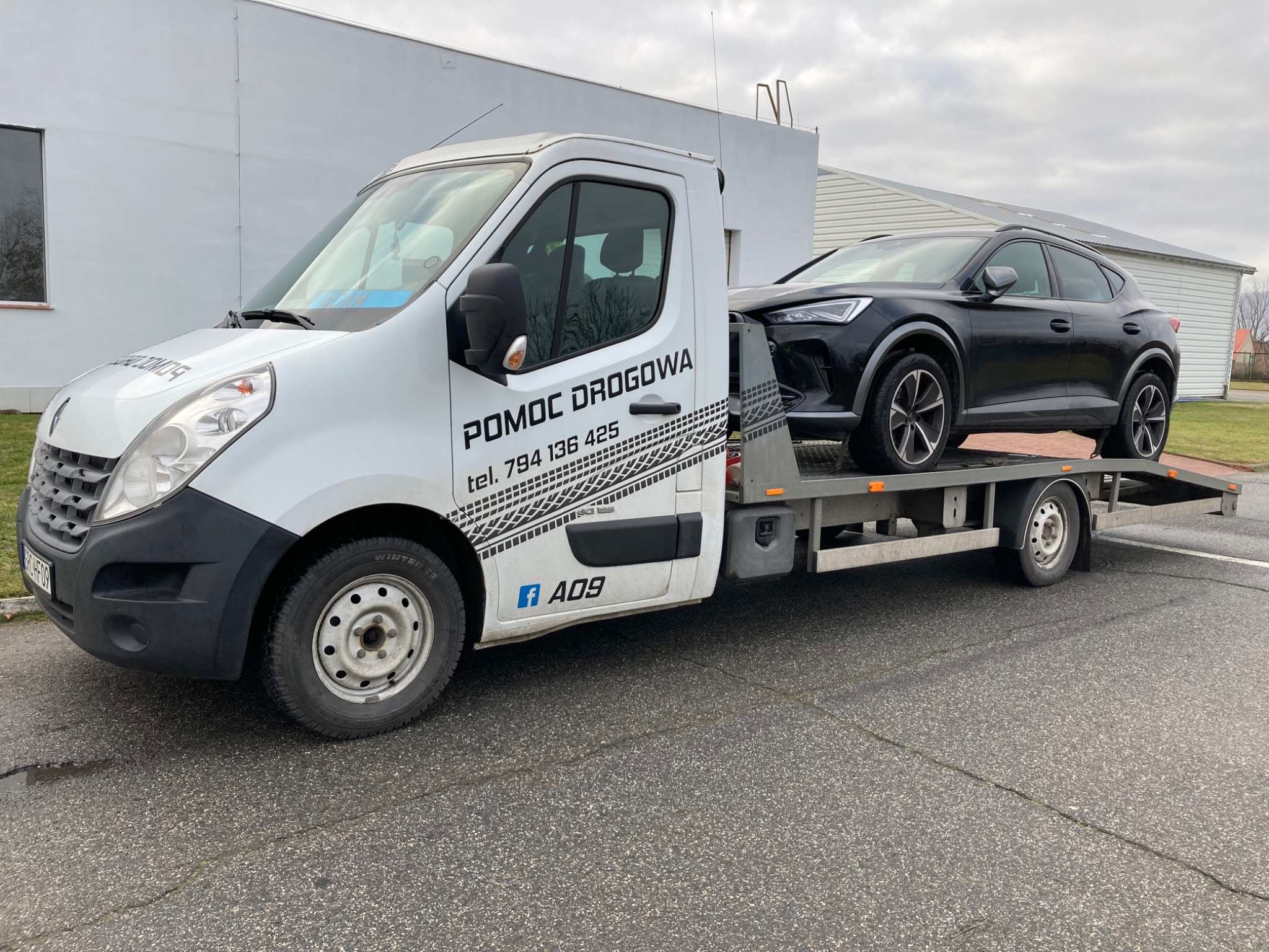 Biała laweta Renault z napisem 'POMOC DROGOWA' i numerem telefonu, przewożąca czarny samochód osobowy Cupra Formentor na platformie, zaparkowana na asfaltowym parkingu przed budynkiem.
