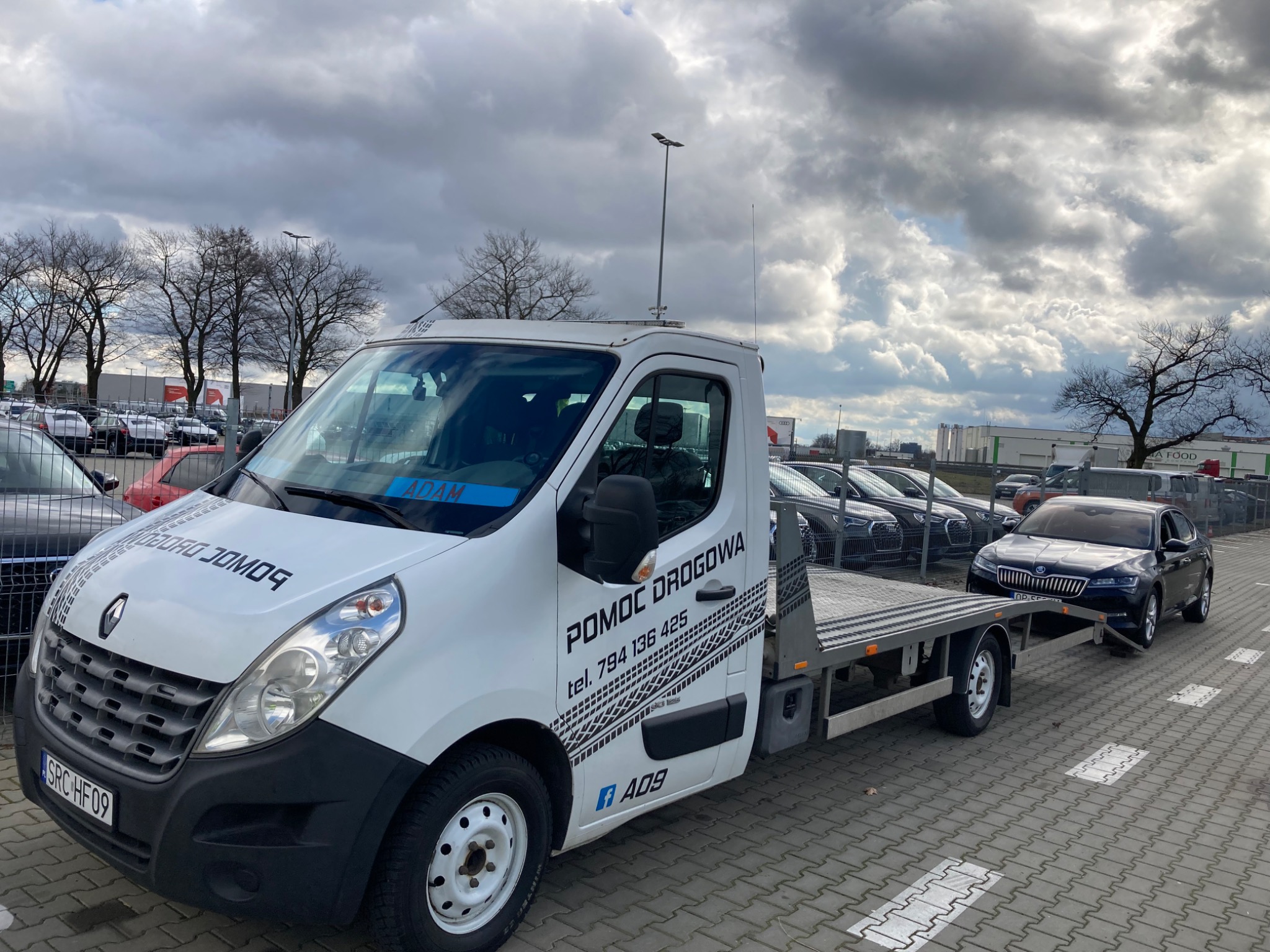 Biała laweta Renault z napisem 'POMOC DROGOWA' i numerem telefonu holuje czarny samochód osobowy na parkingu z szarym brukiem w pochmurny dzień.