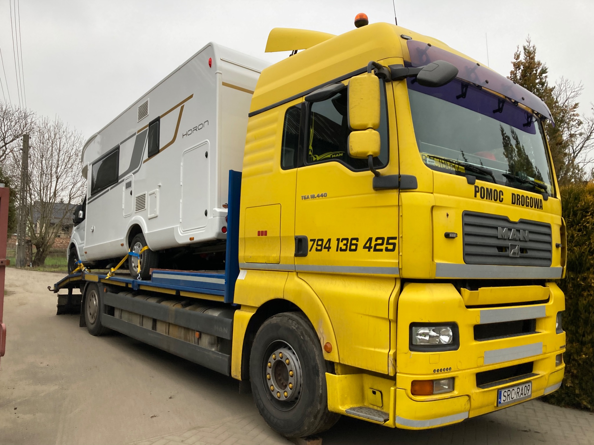 Żółta laweta MAN z napisem 'Pomoc Drogowa' przewozi biały kamper marki Horon, zabezpieczony pasami transportowymi.