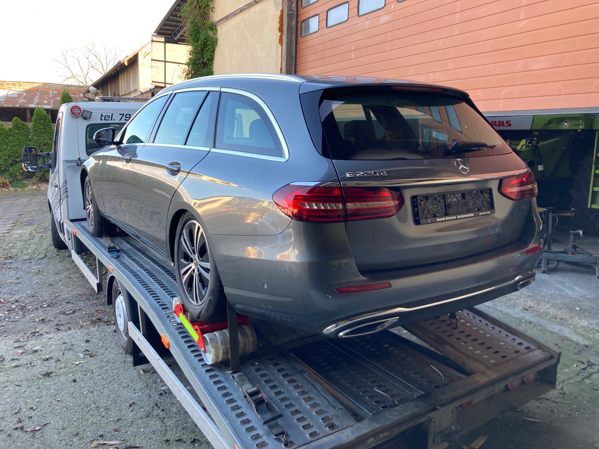 Szary samochód Mercedes-Benz E220d kombi, zabezpieczony pasami transportowymi, na platformie lawety. Widoczny fragment tablicy rejestracyjnej Nord-Ostsee Automobile.