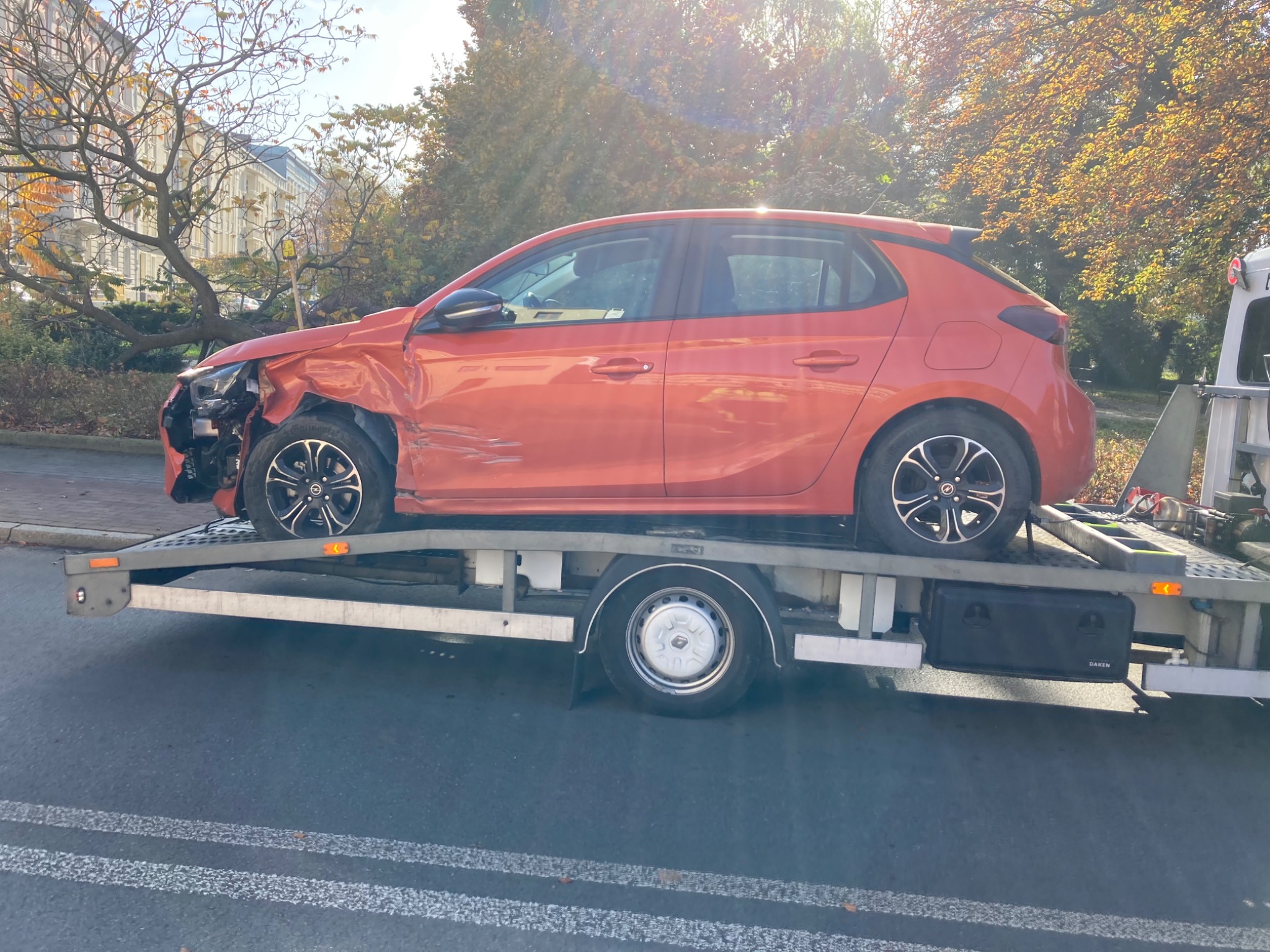 Uszkodzony, pomarańczowy Opel Corsa na platformie lawety, widoczne uszkodzenia przodu pojazdu, jesienne drzewa w tle.