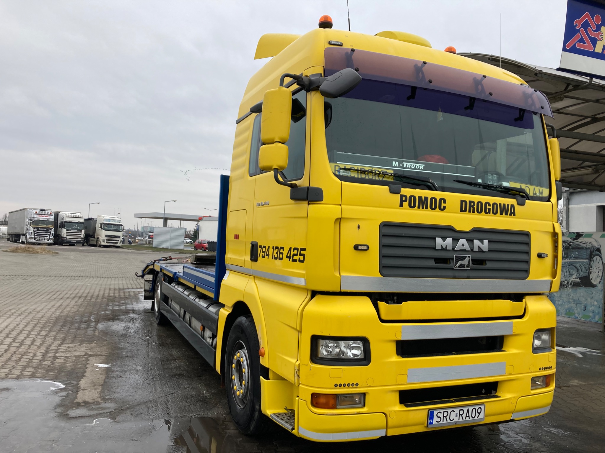 Żółta laweta MAN z napisem 'Pomoc Drogowa' na parkingu z mokrą nawierzchnią, widoczne inne ciężarówki w tle.
