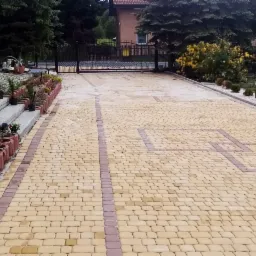 Żółto-brązowa kostka brukowa tworząca wzór na podjeździe, obrzeża z donicami z kwiatami, widok na bramę wjazdową i dom w tle.