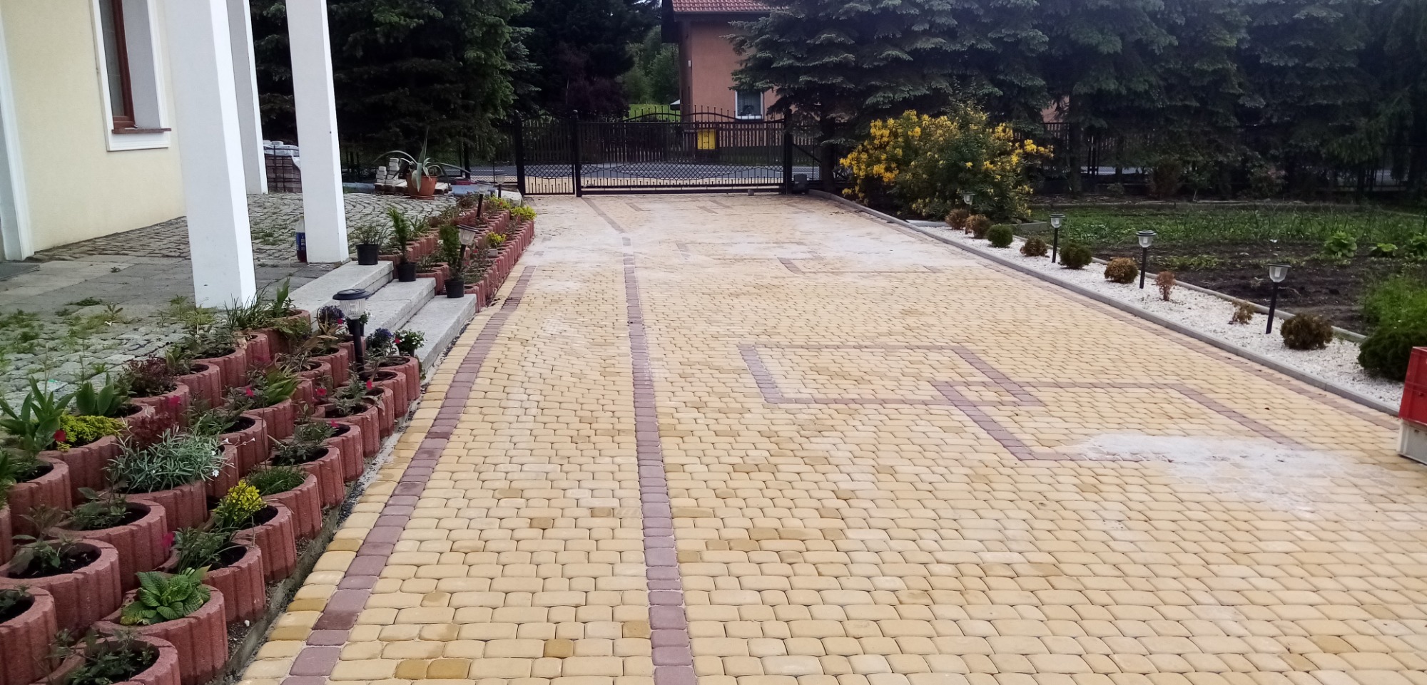 Żółto-brązowa kostka brukowa tworząca wzór na podjeździe, obrzeża z donicami z kwiatami, widok na bramę wjazdową i dom w tle.