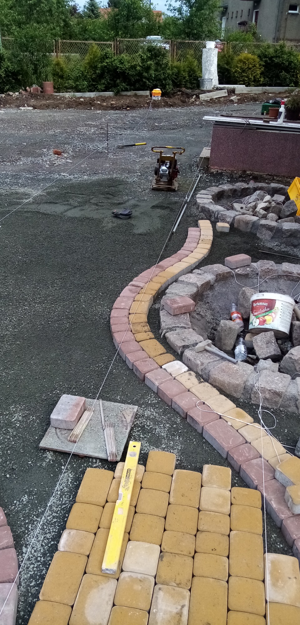 W trakcie prac brukarskich: układanie żółto-czerwonej kostki na podsypce, widoczna poziomica, sznurki wyznaczające linię, zagęszczarka w tle, oraz obrzeże z kamieni.