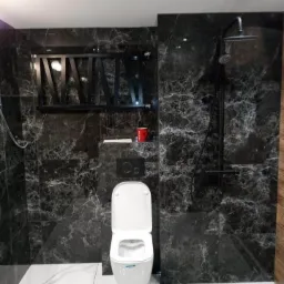 Nowoczesna łazienka w ciemnych marmurkowych płytkach z białymi podłogowymi, wisząca toaleta z czarnym panelem, czarny prysznic deszczownica i dekoracyjna czarna półka.