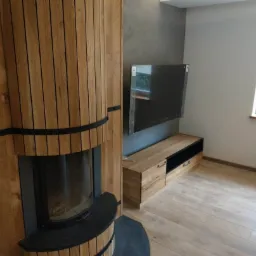Nowoczesny salon z drewnianą obudową kominka z pionowymi listwami, telewizorem na ścianie i drewnianą szafką RTV na parkiecie.