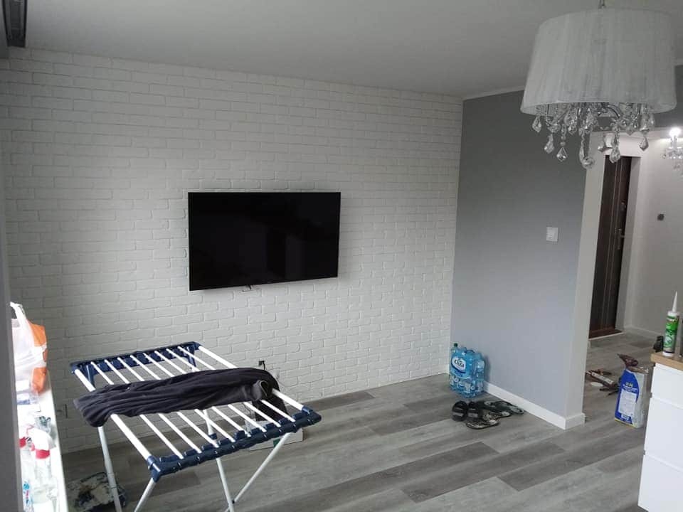 Salon po odświeżeniu: biała ściana z cegły, wiszący telewizor, suszarka z ubraniami, szara ściana, kryształowy żyrandol, panele podłogowe, butelki wody mineralnej i kapcie na podłodze.