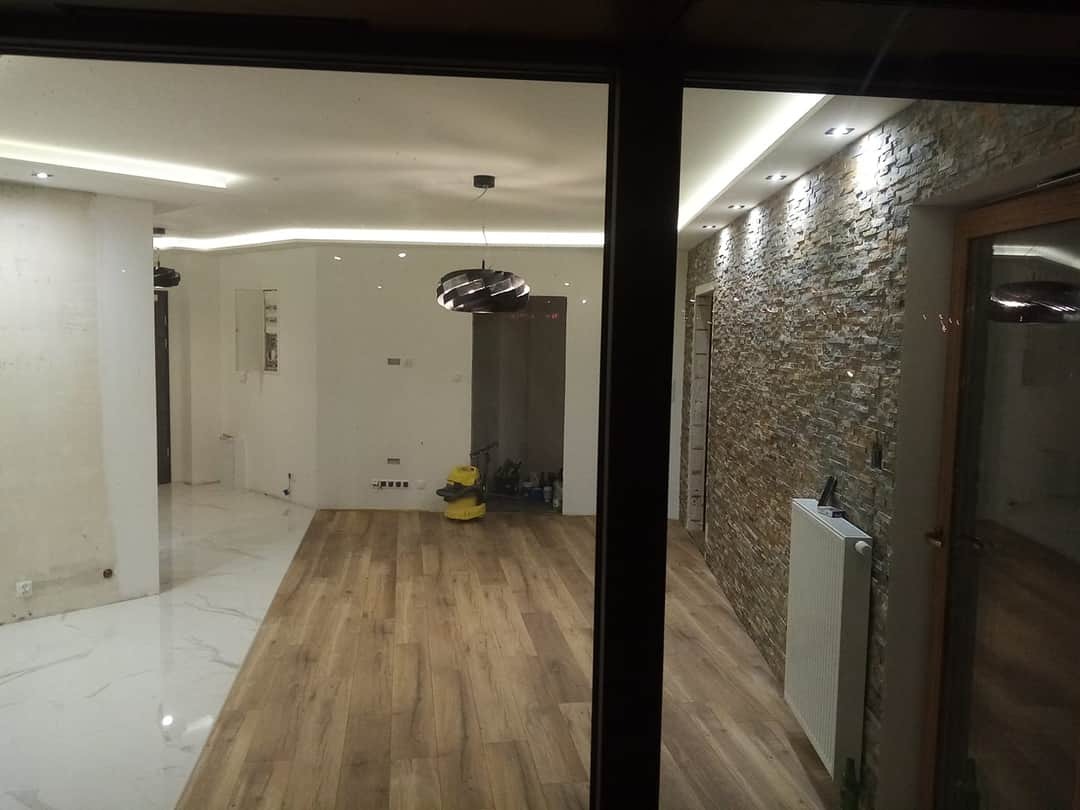 Wnętrze po remoncie: parkiet ułożony, ściana wykończona kamieniem dekoracyjnym, widoczny grzejnik i lampa wisząca, część pomieszczenia z marmurową podłogą w trakcie prac wykończeniowych.