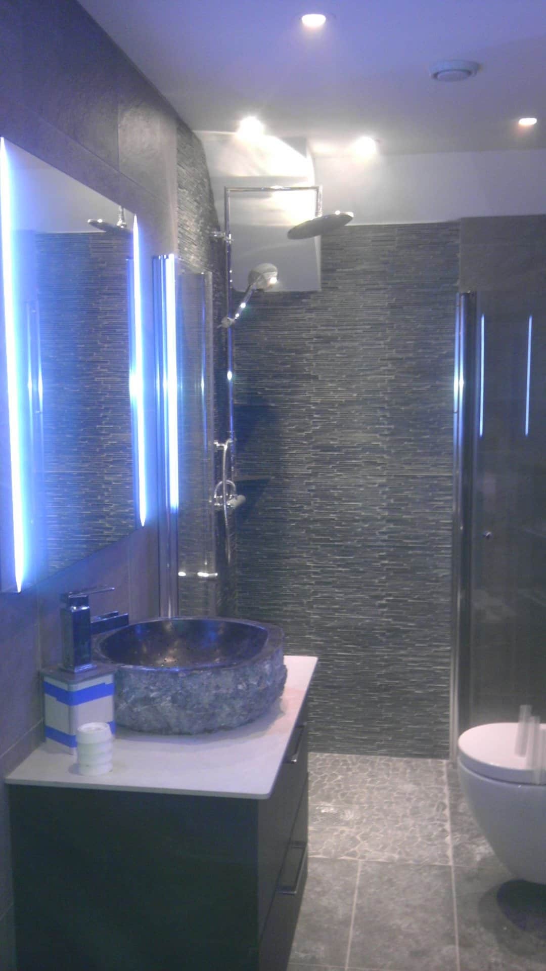 Nowoczesna łazienka z kamienną umywalką, podświetlanym lustrem, kabiną prysznicową i toaletą w szarych odcieniach, wykończona płytkami imitującymi kamień.