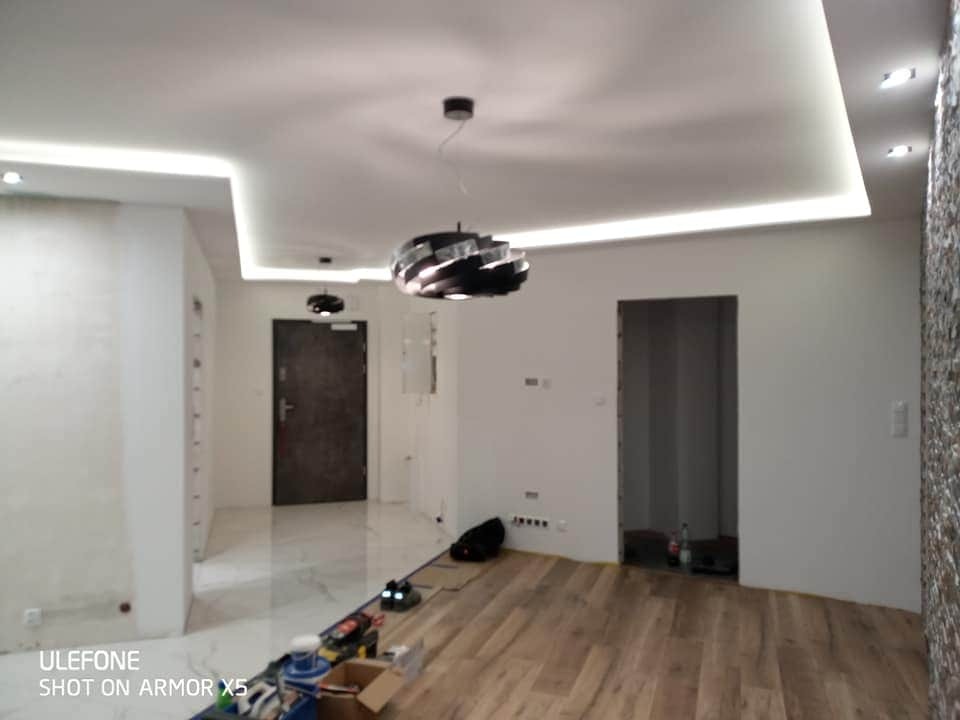 Wnętrze mieszkania w trakcie prac wykończeniowych: drewniana podłoga, pomalowane na biało ściany, dekoracyjny kamień na ścianie, podwieszany sufit z oświetleniem LED, nowoczesne lampy wiszące...