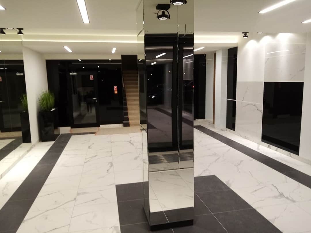 Nowoczesne wnętrze holu z marmurową podłogą w odcieniach bieli i szarości, lustrzaną kolumną i czarnymi akcentami na ścianach, oświetlone ledowymi lampami sufitowymi.