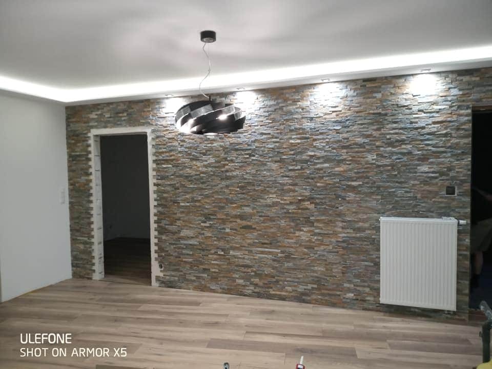 Ściana wyłożona dekoracyjnym kamieniem w odcieniach brązu i szarości, oświetlona ledowym pasem na suficie podwieszanym, z wiszącą lampą w kształcie spirali i grzejnikiem na ścianie, widoczna...