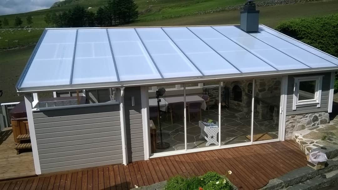 Zadaszony taras z przeszklonymi ścianami, kamienną podłogą i widokiem na zielone wzgórza; widoczny fragment jacuzzi i stół z krzesłami.