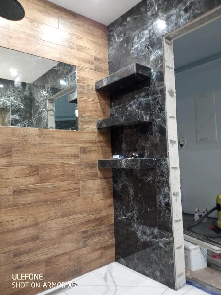 Wykończenie łazienki z drewnopodobnymi panelami ściennymi, czarnym marmurem i półkami z tego samego materiału, widoczne niedokończone obramowanie drzwi.