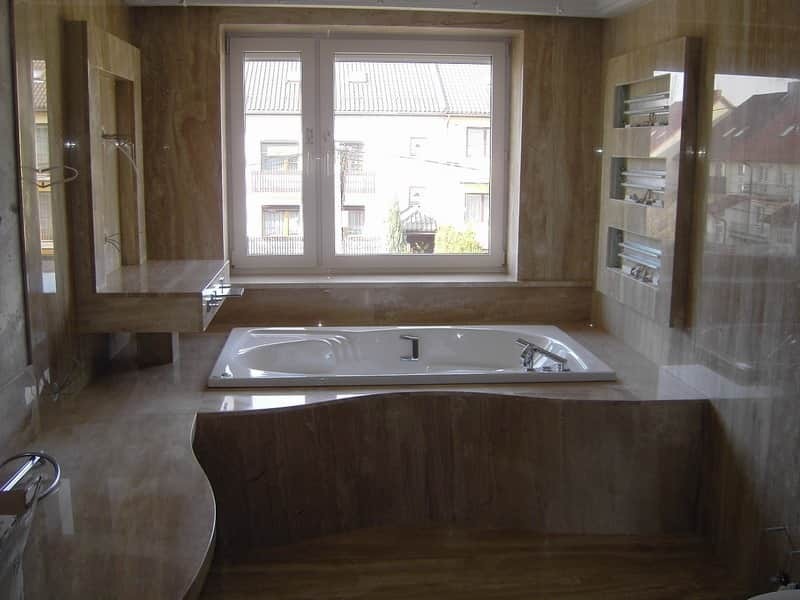 Wykończona łazienka z beżowymi marmurkowymi płytkami, białą wanną pod oknem i zabudowaną umywalką z lustrem.