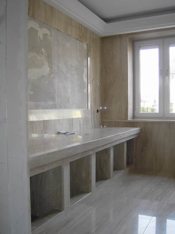 Łazienka w trakcie wykańczania: marmurowe ściany i podłoga, blat z wnękami pod umywalkę, ściana przygotowana pod płytki, widoczne okno z naturalnym światłem.