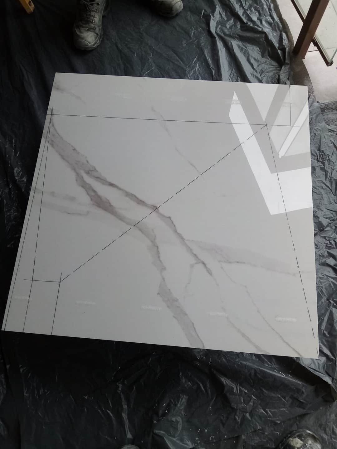 Płytka ceramiczna imitująca marmur z narysowanymi liniami cięcia, leżąca na czarnej folii ochronnej, widoczne buty robocze w górnym rogu.