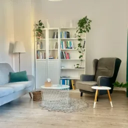 Jasne, przytulne wnętrze gabinetu z szarym kanapą i fotelem, białą biblioteczką z książkami i roślinami, oraz okrągłym stolikiem kawowym na puszystym dywanie.