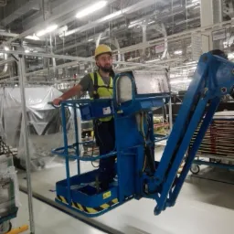Pracownik w żółtym kasku i kamizelce odblaskowej na podnośniku koszowym w hali produkcyjnej, widoczne instalacje pod sufitem.