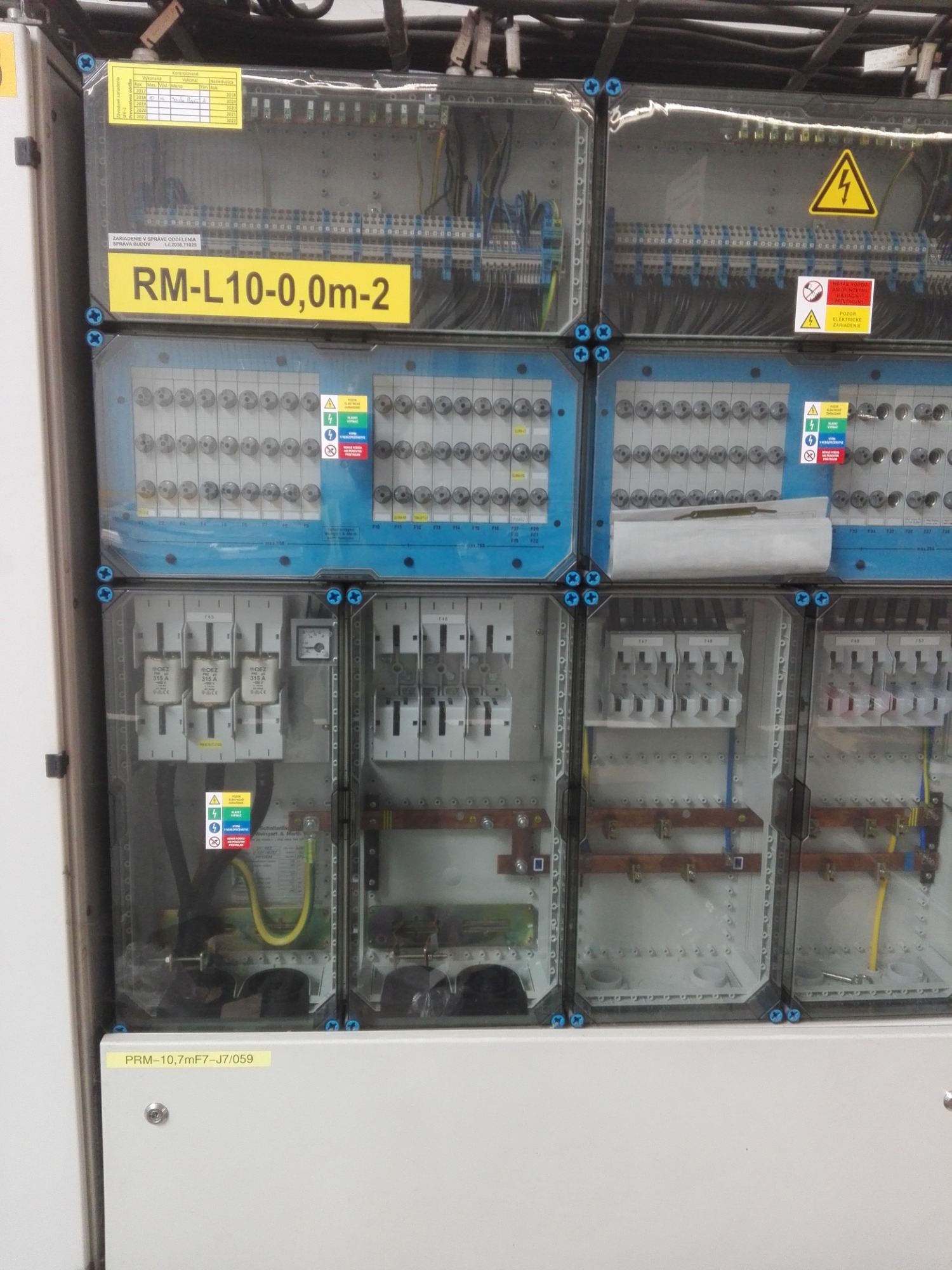 Rozbudowana tablica elektryczna z bezpiecznikami i oznaczeniami, widoczne kable, elementy zabezpieczeń i ostrzeżenia o napięciu.